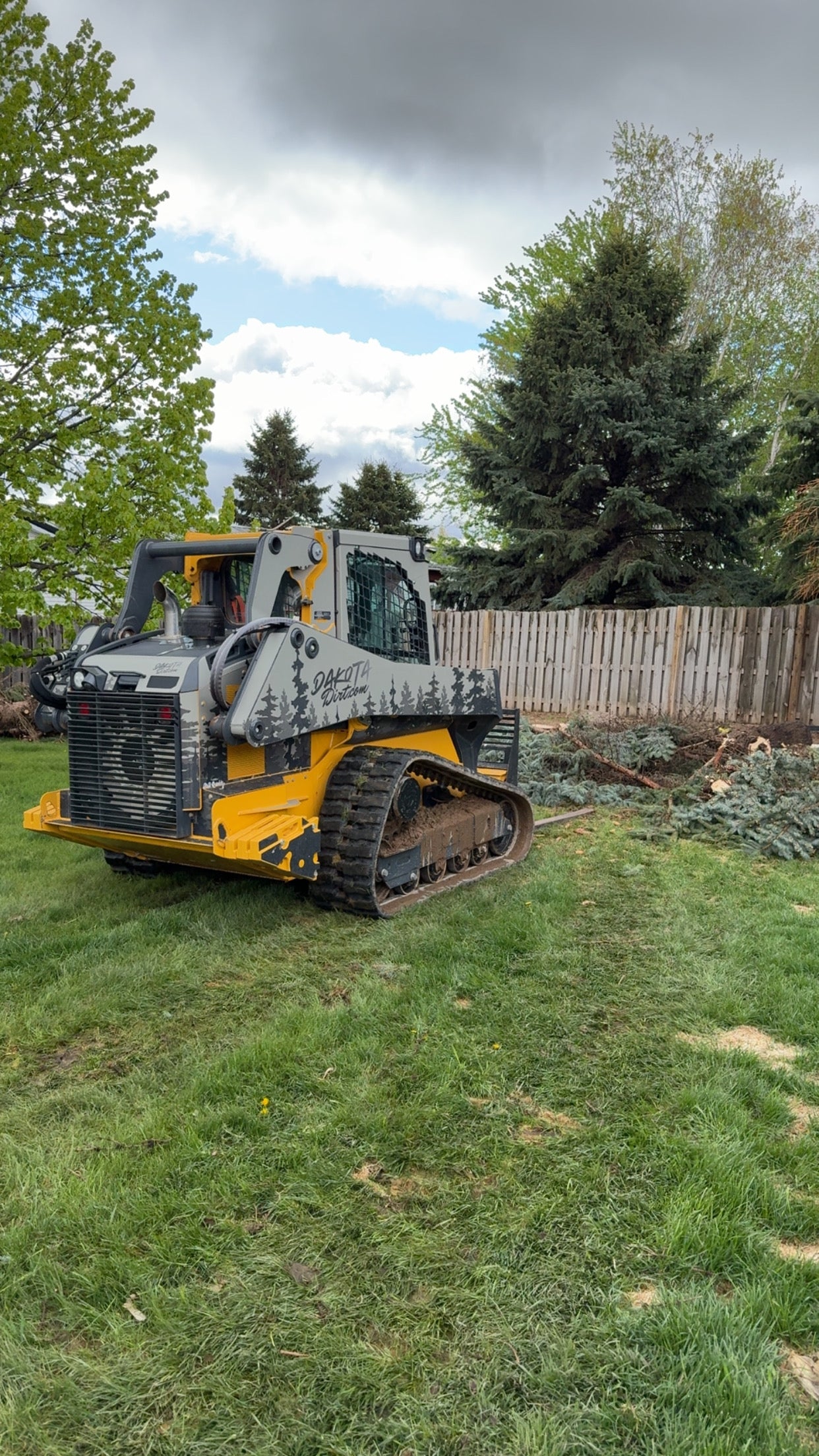 Large construction vehicle on grass with trees and a wooden fence in the background